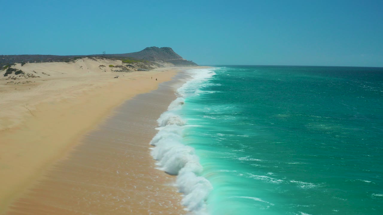 Opening frame of La Laguna Beach in Los Cabos, Mexico, with turquoise ocean on the right, white surf reaching the sand, dunes and a distant headland under clear sky, and no visible people.