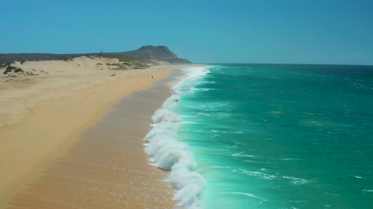La Laguna Beach shoreline one second in, with a wider band of white foam pushing onto tan sand while the turquoise sea fills the right side and the headland remains in the distance, no visible people.