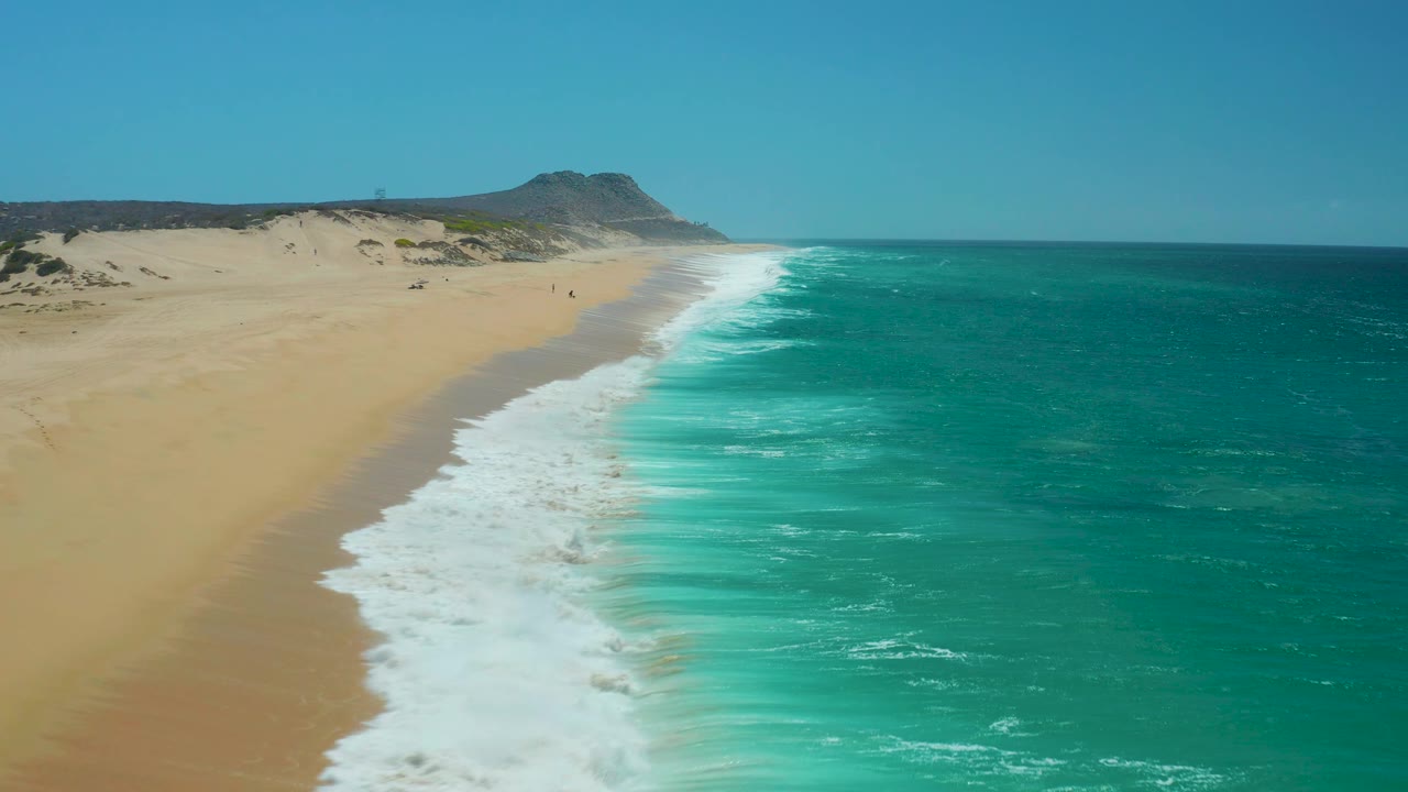 At 0:02, the breaking surf forms a broad white strip between the turquoise ocean and sandy La Laguna Beach, with low dunes and a rocky headland across the background, no visible people.