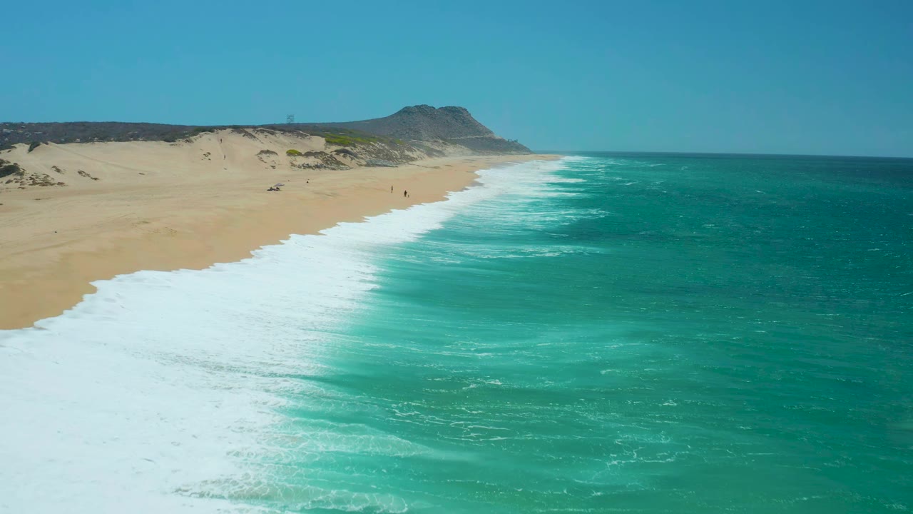 Aerial view at La Laguna Beach Los Cabos, Mexico, where a broad white surf band pushes farther up the sand beside turquoise water, with dunes and a headland in the background and no visible people.