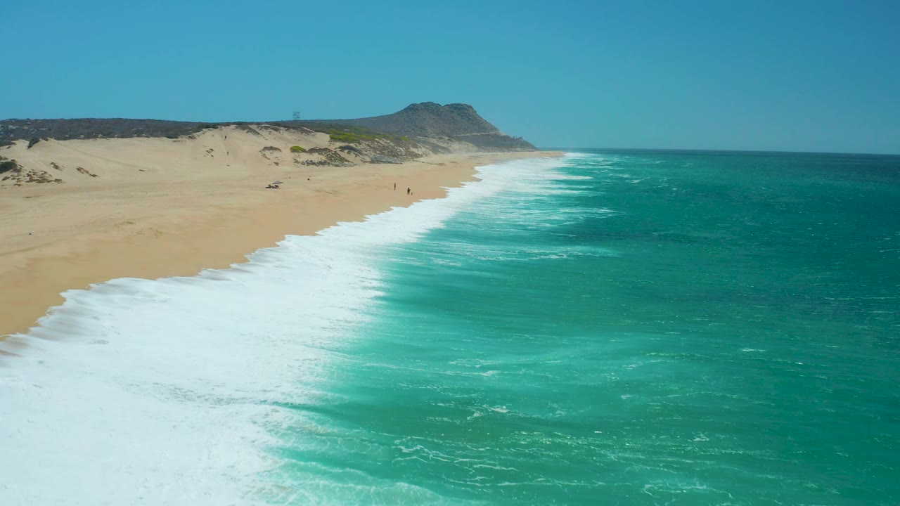 La Laguna Beach Los Cabos, Mexico, shown from above with foamy turquoise surf curving along the right edge of open sand, low dunes behind the beach, and no visible people.