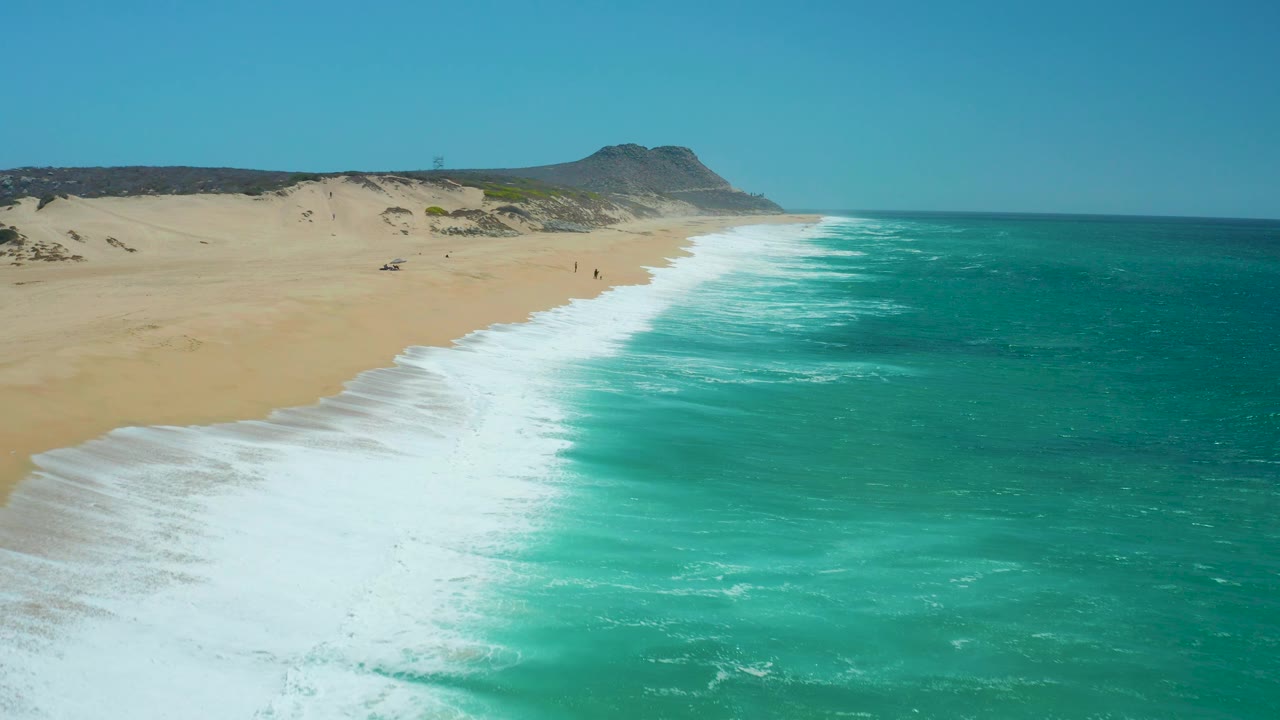 Aerial frame from La Laguna Beach Los Cabos, Mexico, with white foam washing over wet sand in the foreground, deeper turquoise water offshore, dunes inland, and no visible people.