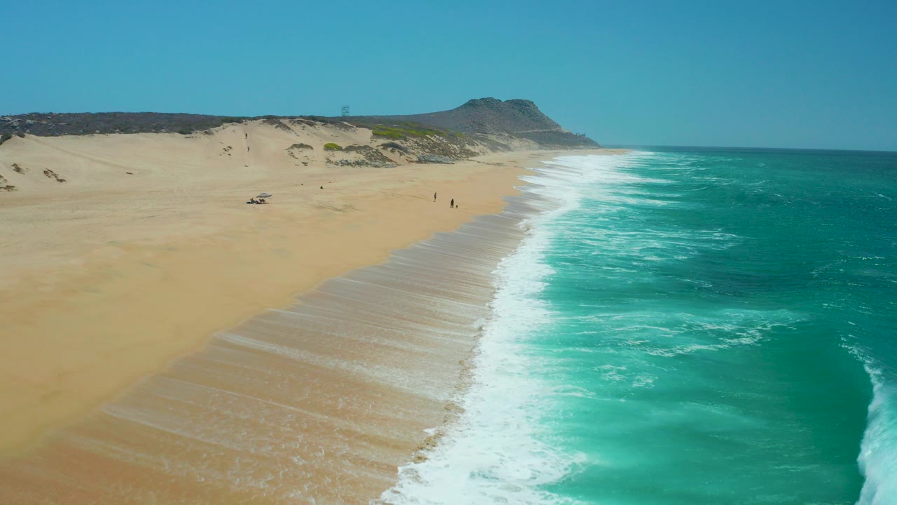 Aerial view over La Laguna Beach in Los Cabos, Mexico, with broad sand on the left, turquoise surf on the right, low dunes beyond the beach, and tiny people visible near the shoreline.