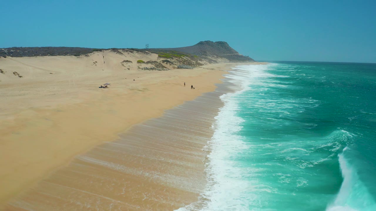 Aerial view of La Laguna Beach with a stronger wave pattern on the right, pale sand on the left, dune ridges beyond the shore, and tiny beachgoers visible near the center distance.