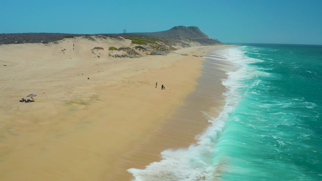 High aerial frame over La Laguna Beach Los Cabos, Mexico, with turquoise surf on the right, broad tan sand on the left, dunes beyond the beach, and small distant people near the water.