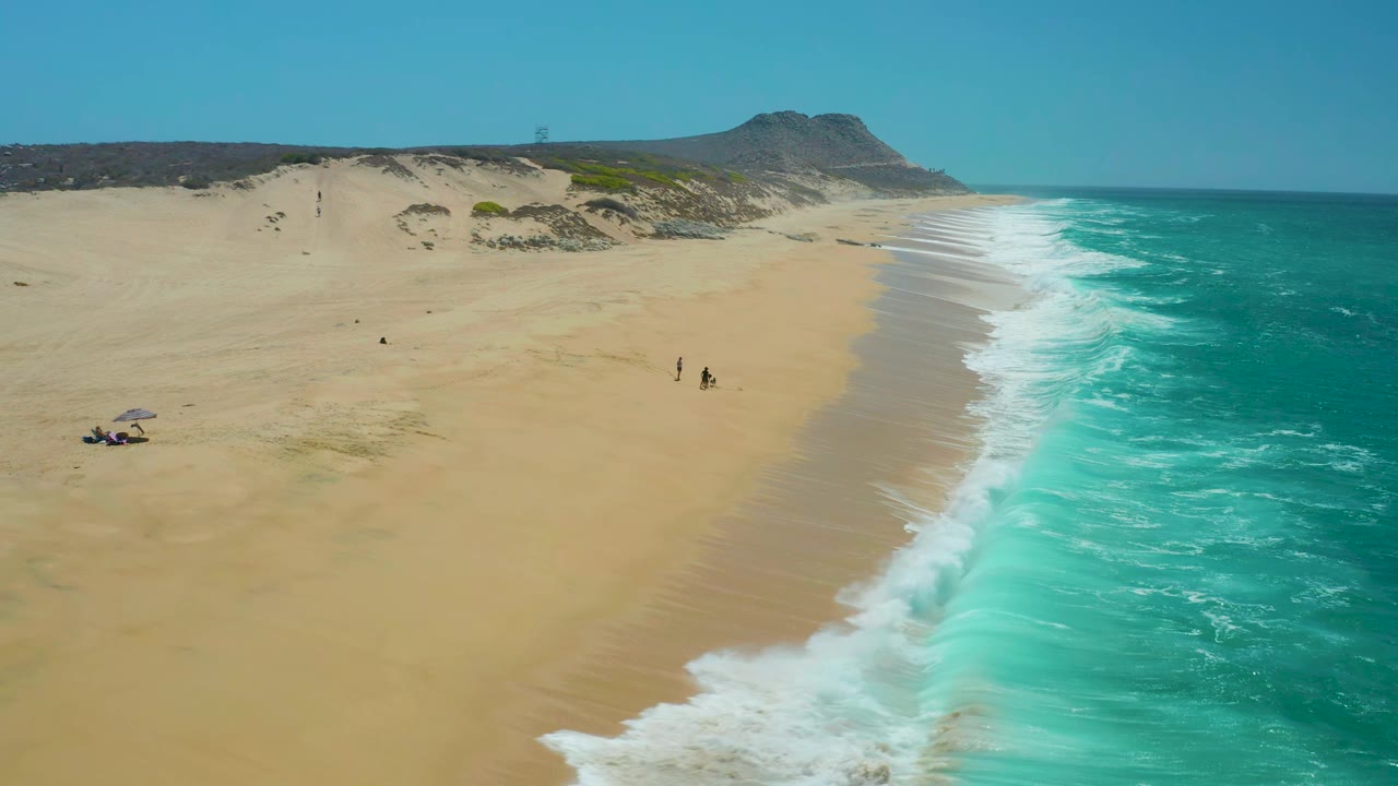 Drone view along a sandy shoreline with waves and distant beachgoers.