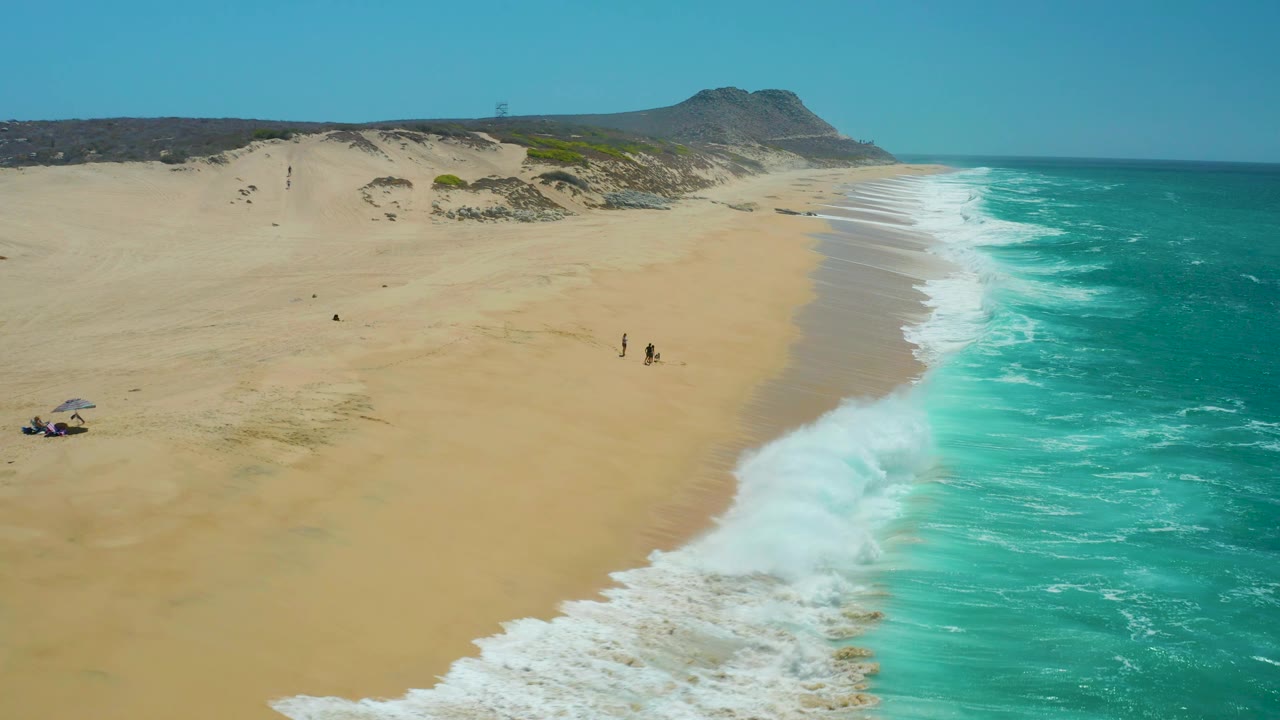 La Laguna Beach Los Cabos, Mexico appears from above as a broad white breaker reaches farther onto the tan sand, with dunes behind and small people still visible near the shoreline.