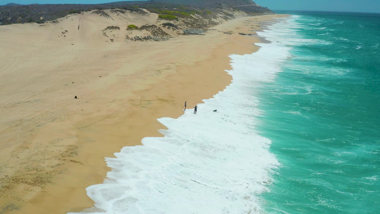 Aerial view at La Laguna Beach Los Cabos with a broad sandy beach on the left, turquoise ocean on the right, white surf sweeping diagonally, and small people near the waterline.