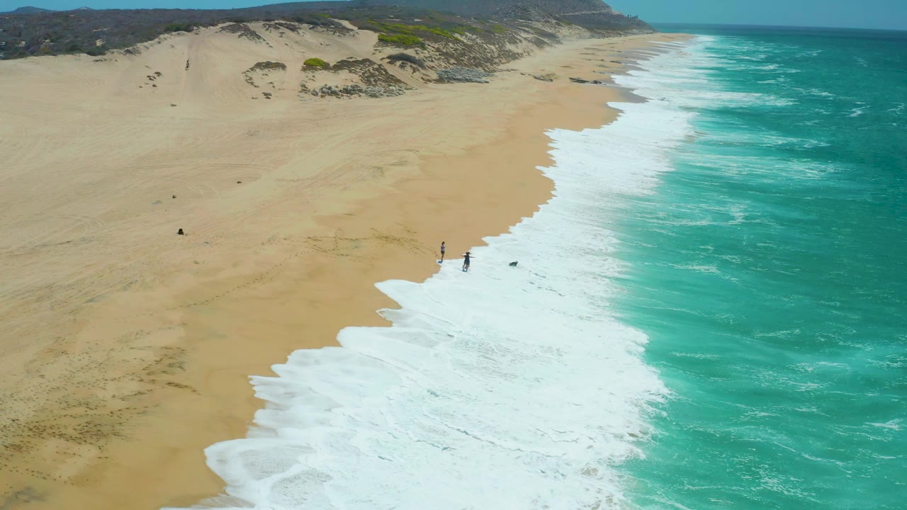 Wide aerial shoreline with foam pushing farther up a sandy beach beside turquoise water.