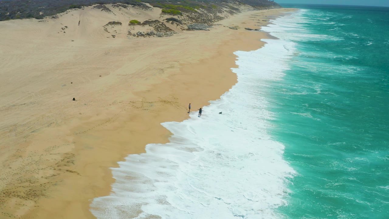 At 0:02, La Laguna Beach Los Cabos is seen from above with a diagonal band of white surf, broad empty sand, dune areas inland, and a few tiny people near the wash.