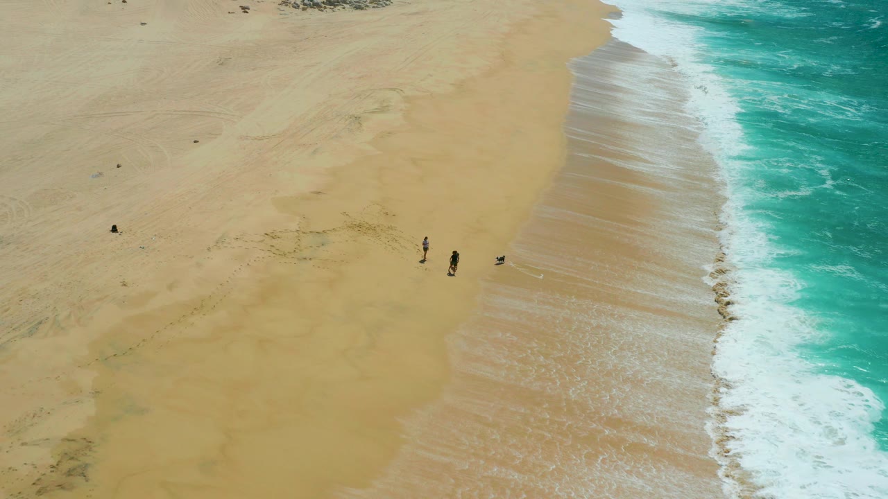 Overhead frame from La Laguna Beach in Los Cabos showing broad tan sand, turquoise surf on the right, and several people walking near the shoreline.