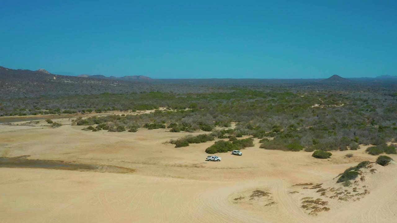 Aerial frame at 0:01 with the two SUVs slightly separated on the sand, backed by green-brown scrubland, mountain ridges, a flat horizon, and no visible people.