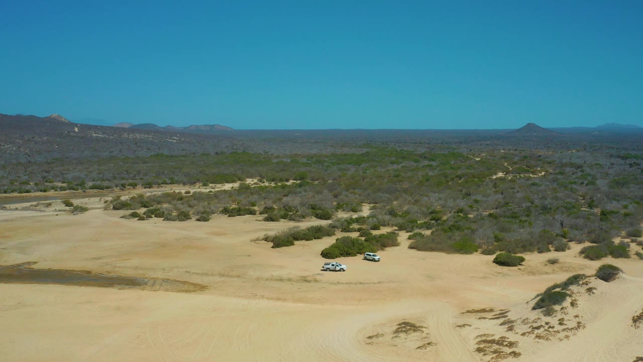 At 0:02, the aerial view shows the SUVs near the lower sandy area, with a darker vegetated desert plain behind them, low mountains on both sides, and no visible people.