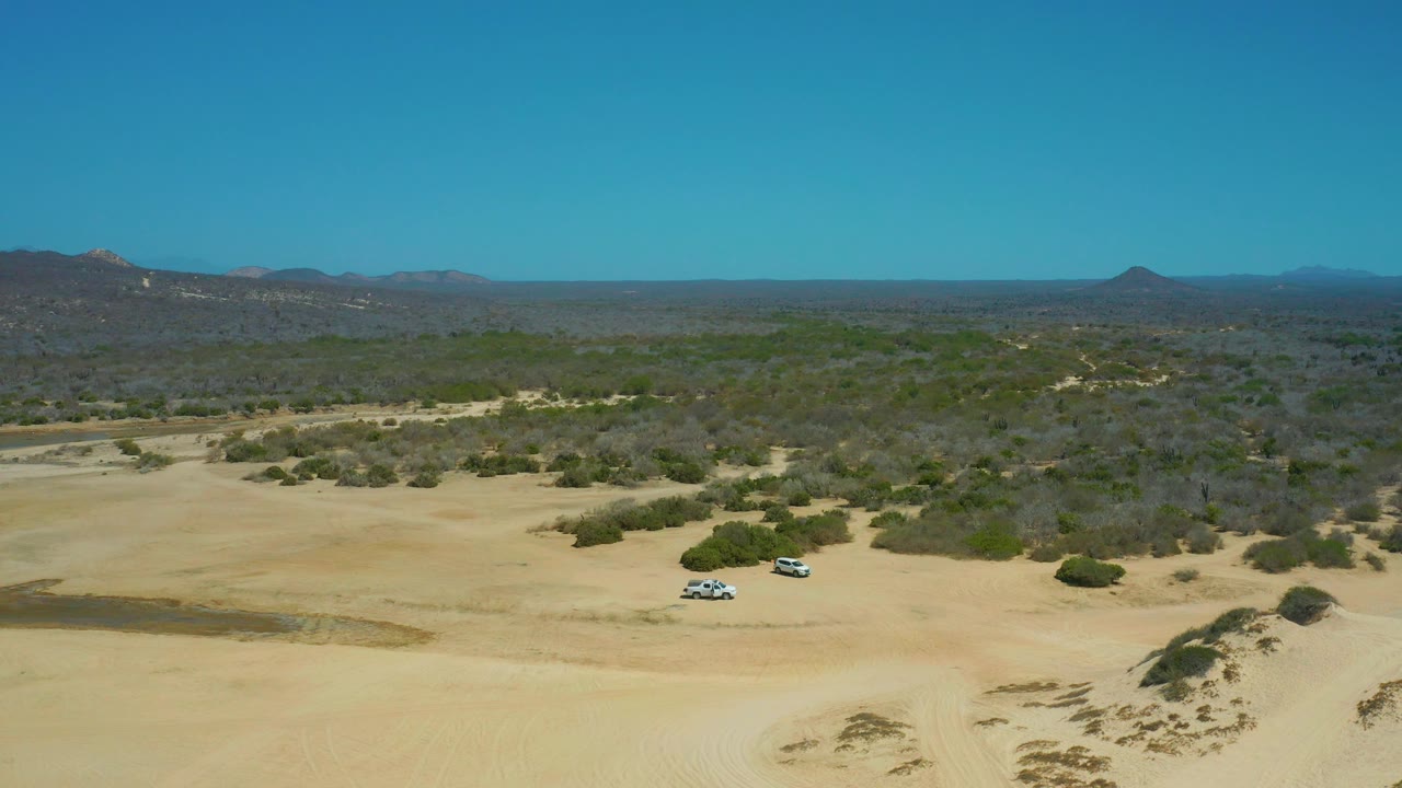 Mid-clip aerial frame from La Laguna Beach Los Cabos with two SUVs on an open sandy clearing, scattered shrubs, broad mountain silhouettes, bright daylight, and no visible people.