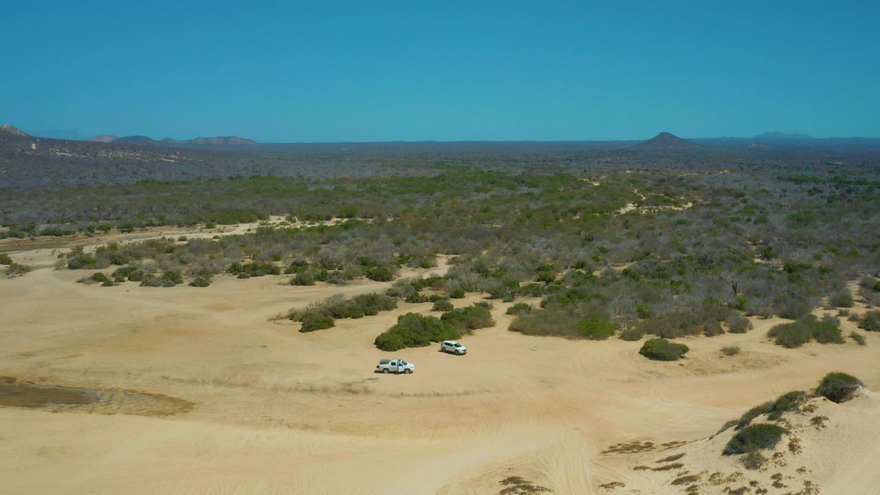 Aerial frame at La Laguna Beach Los Cabos, Mexico showing two light-colored SUVs on pale sand, sparse shrubs, distant hills, clear blue sky, and no visible people.
