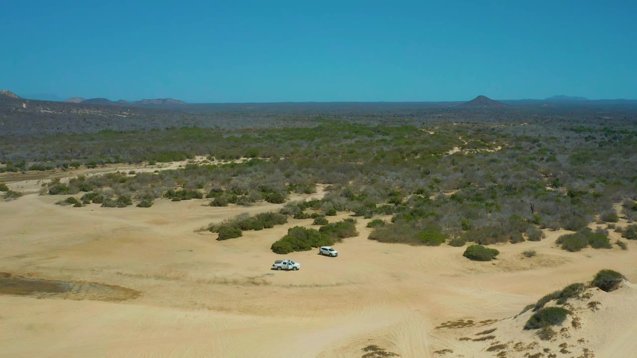 Two SUVs crossing open sand with scrubland and low hills beyond.