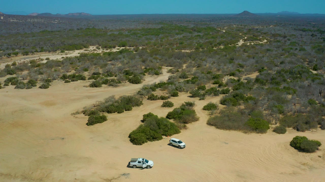 Aerial view at La Laguna Beach Los Cabos, Mexico, showing two white vehicles on open pale sand below green-brown scrubland, distant hills, blue sky, and no visible people.