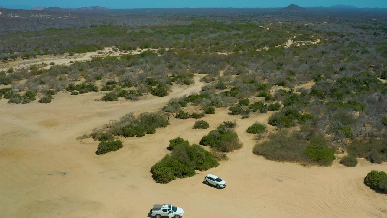 Aerial view over La Laguna Beach Los Cabos, Mexico, where two white vehicles sit near the lower edge of a sandy clearing with scrubland and distant hills behind them, no visible people.
