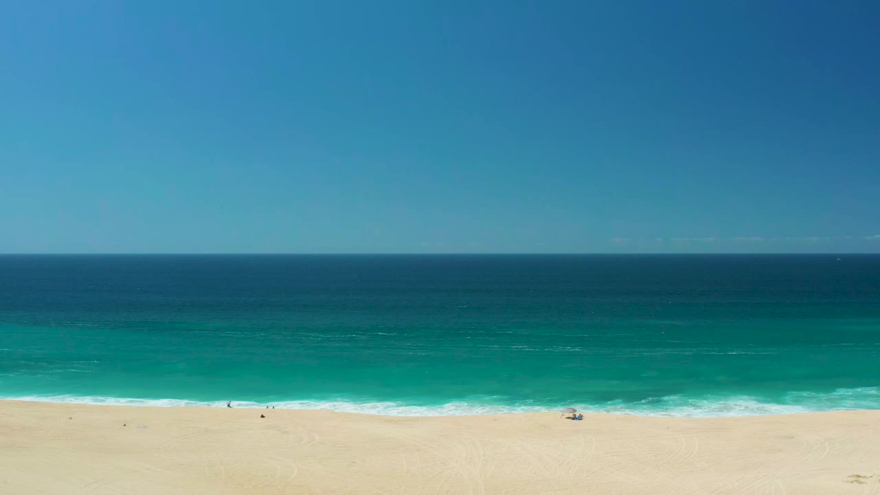 Wide aerial view of La Laguna Beach Los Cabos with pale sand at the bottom, turquoise shore break, deep blue ocean, clear sky, and tiny beachgoers near the waterline.