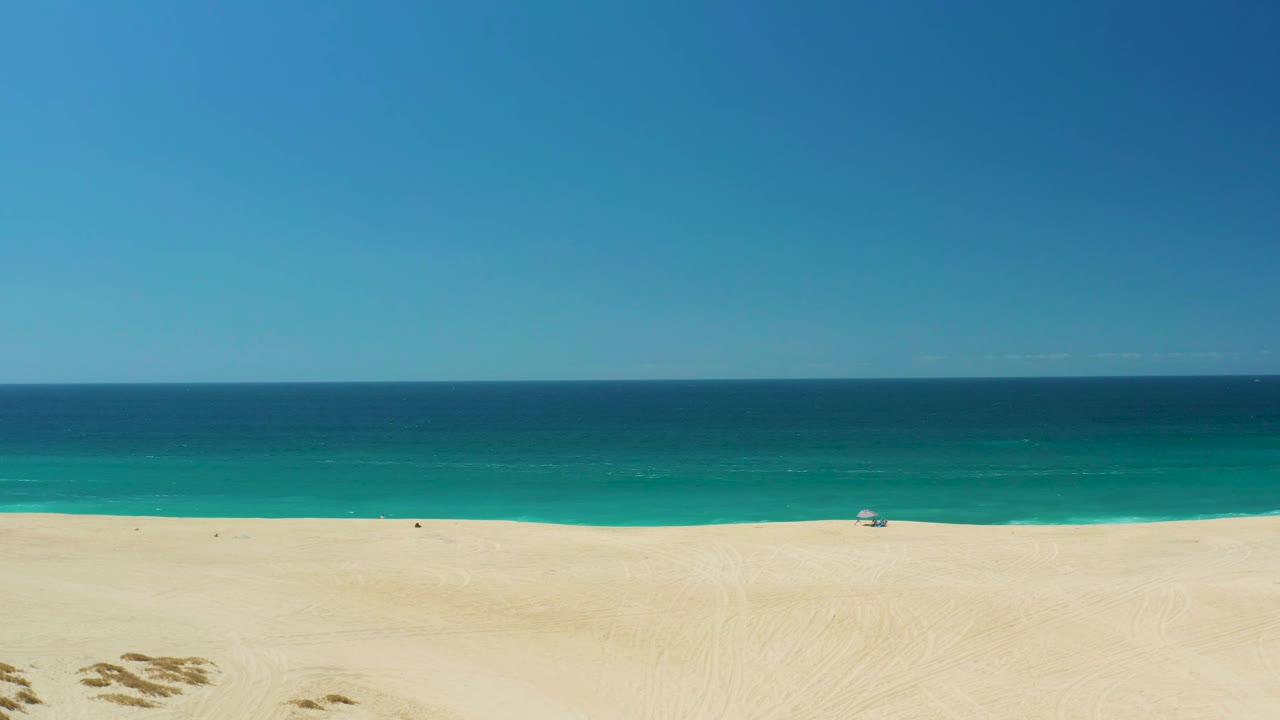 Aerial view at 0:01 over La Laguna Beach Los Cabos, showing open sand, a narrow surf line, teal water, deep blue ocean, and no visible people.