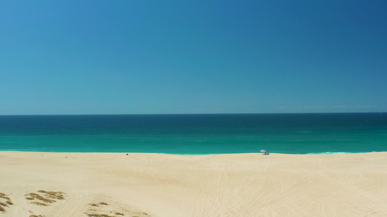 La Laguna Beach Los Cabos aerial frame with pale sand and low scrub patches in the foreground, turquoise water beyond, clear sky above, and no visible people.
