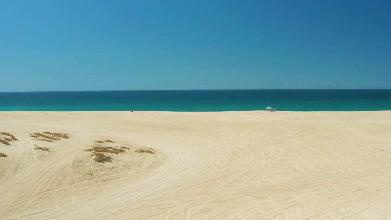 Wide frame of La Laguna Beach in Los Cabos with pale sand in front, low dune patches to the left, blue ocean on the horizon, and no visible people.