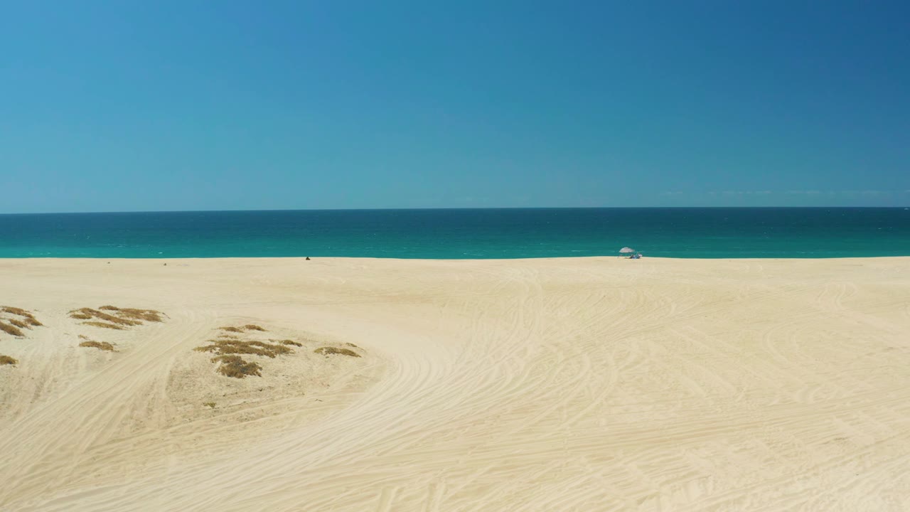 La Laguna Beach view with dune mounds slightly nearer in the sand field, a narrow turquoise shoreline band, deeper blue water beyond, and no visible people.