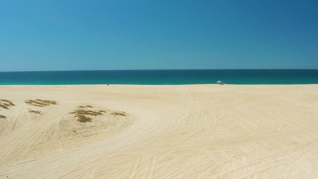 Wide La Laguna Beach frame showing tracked sand in the foreground, darker dune patches near center left, blue ocean across the background, and no visible people.