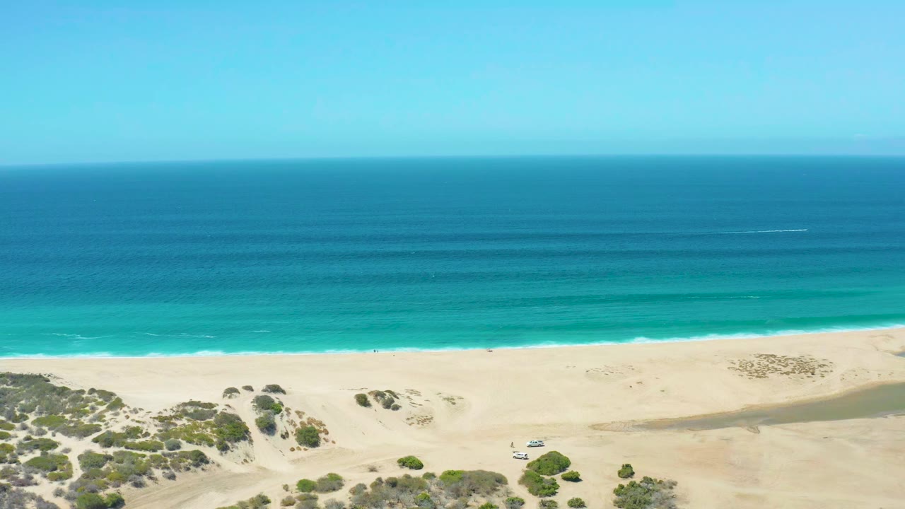 Aerial view over La Laguna Beach with a broad blue ocean horizon, thin surf line, pale sand, dune patches, a small vehicle near the shore, and no visible people.