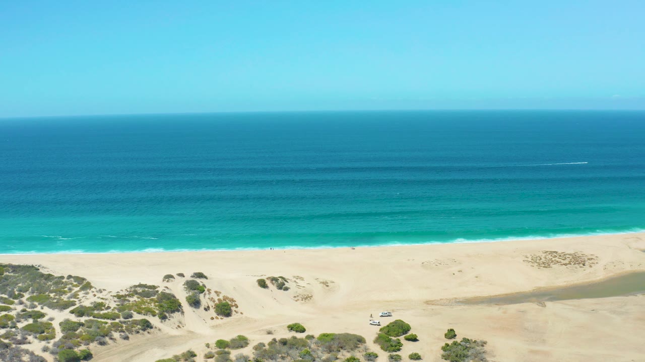 Drone-height frame of La Laguna Beach showing the ocean horizon, blue-green water bands, smooth sand, dune vegetation, a tiny beach vehicle, and no visible people.