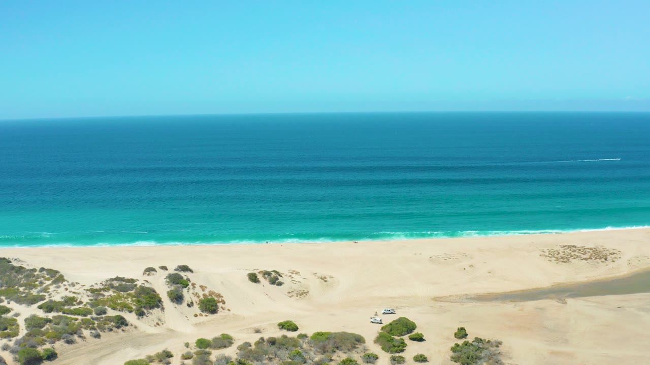 Aerial frame from La Laguna Beach Los Cabos showing a white vehicle on open sand below dunes and shrubs, with turquoise water, darker ocean bands, clear sky, and no visible people.
