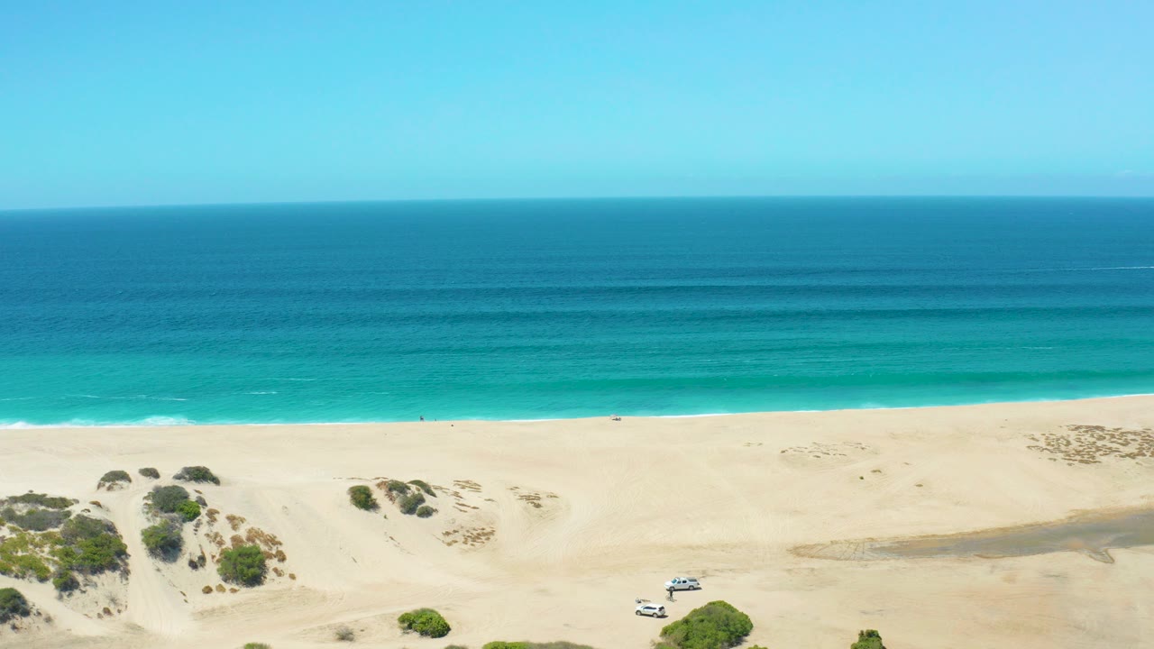 Opening frame of La Laguna Beach Los Cabos with pale dunes, sparse bushes, a white SUV near the lower sand, layered blue ocean, clear sky, and no visible people.