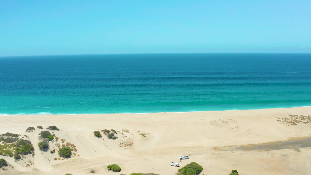 At 0:01, the La Laguna Beach Los Cabos frame shows the white SUV slightly farther left on the sand, with low dune shrubs below a calm turquoise shoreline and no visible people.