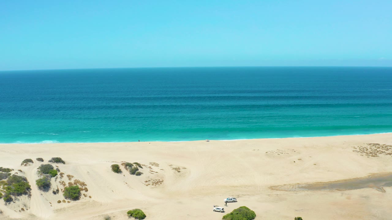 At 0:02, the SUV sits closer to the center-left sand at La Laguna Beach Los Cabos, with brush clusters in the dunes, soft wave lines offshore, and no visible people.
