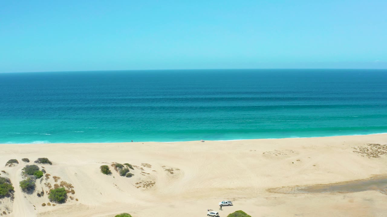 At 0:03, the white SUV has progressed left along the dune edge at La Laguna Beach Los Cabos, while the surf line runs horizontally beneath a wide blue ocean and no visible people appear.