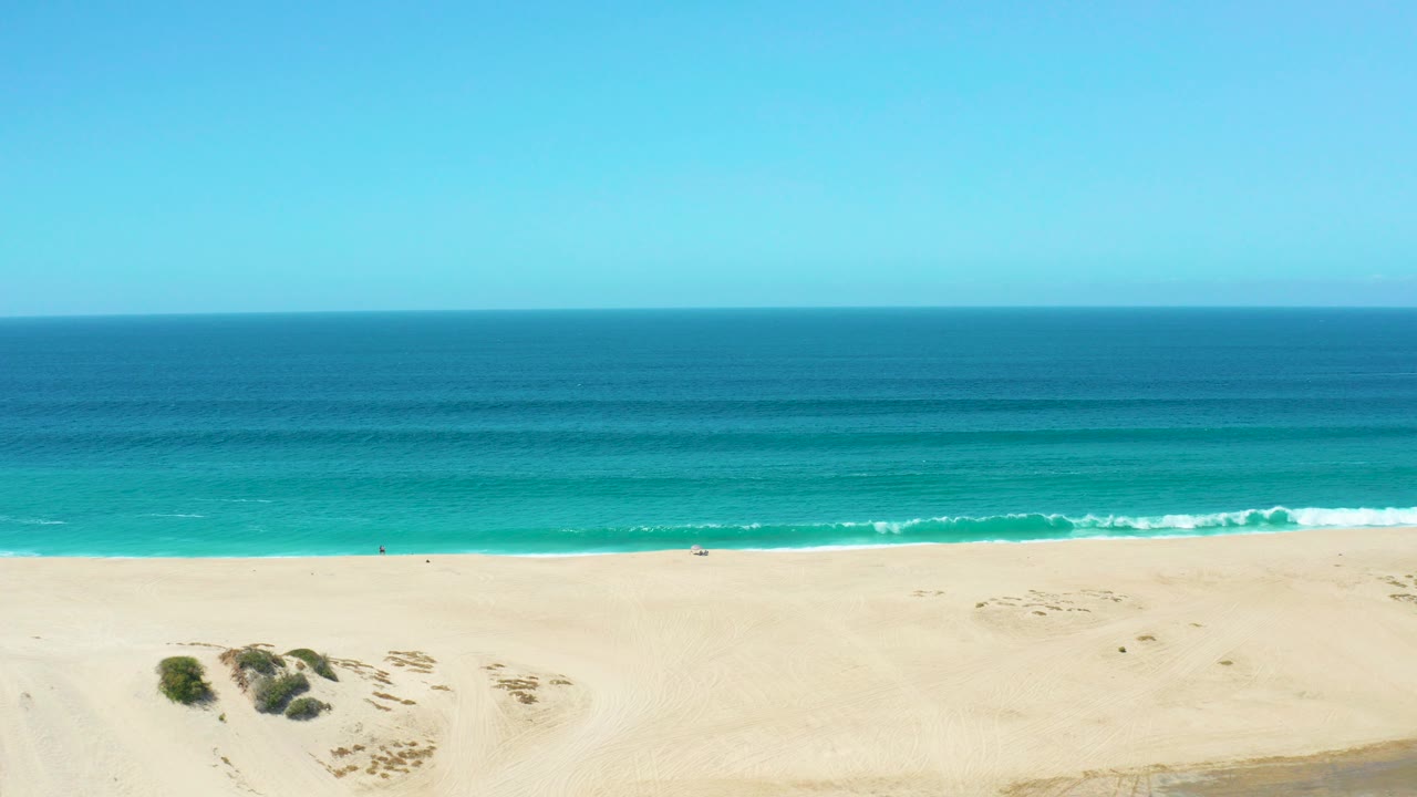 Elevated view of La Laguna Beach Los Cabos with pale sand below, turquoise surf near shore, deep blue ocean beyond, and tiny distant beachgoers visible.