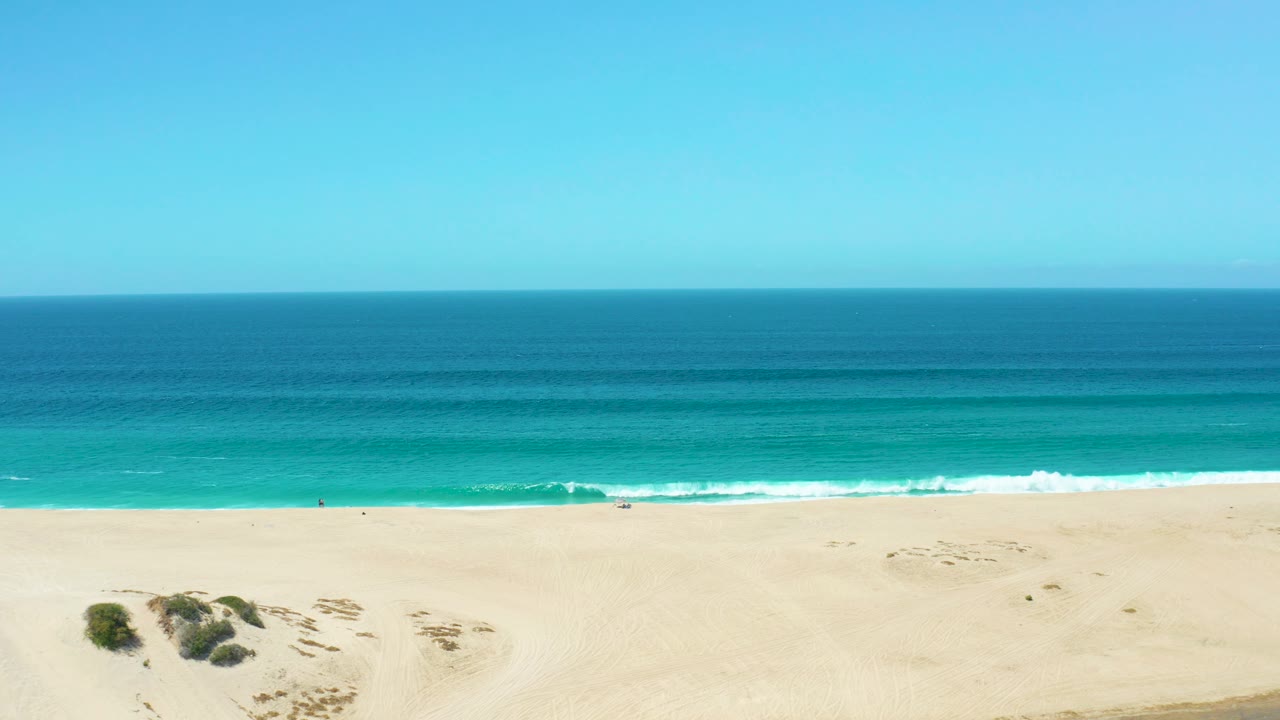 The La Laguna Beach Los Cabos shoreline fills the lower frame while turquoise water and darker offshore bands stretch toward a clear horizon, with tiny distant people on the sand.