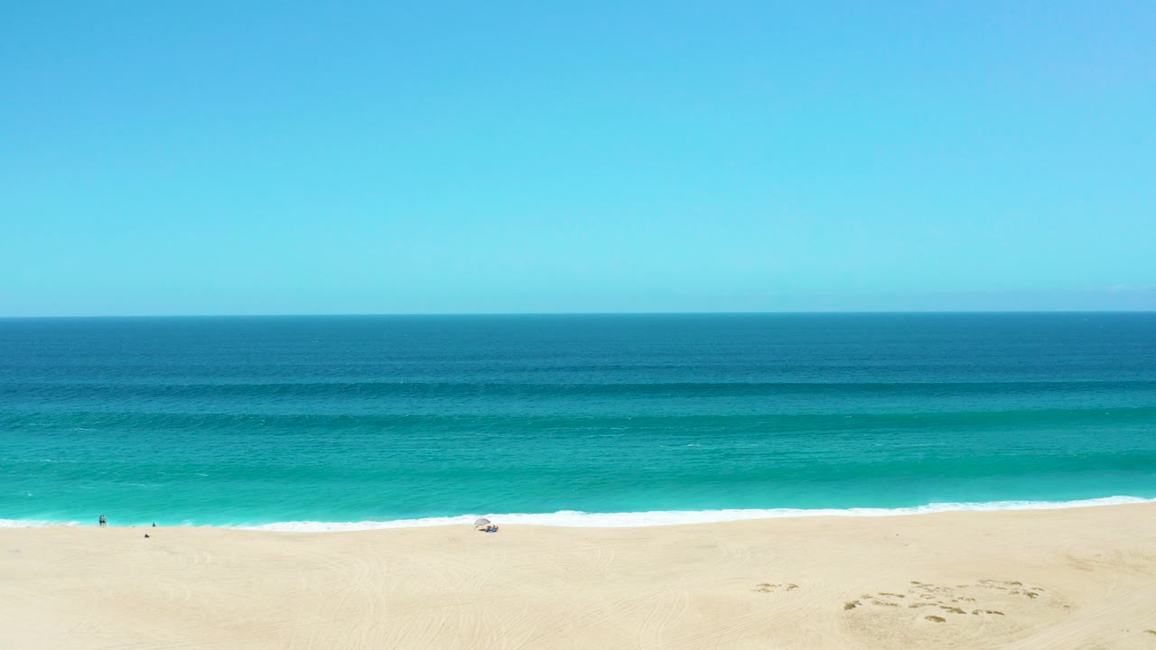 Wide elevated view of La Laguna Beach Los Cabos with pale sand at the bottom, turquoise surf bands, a straight ocean horizon, and tiny people visible near the shoreline.