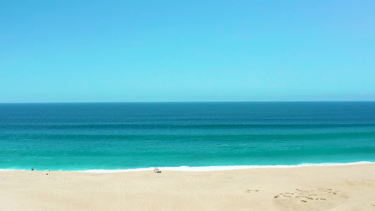 Turquoise waves approaching a sandy beach under clear sky.