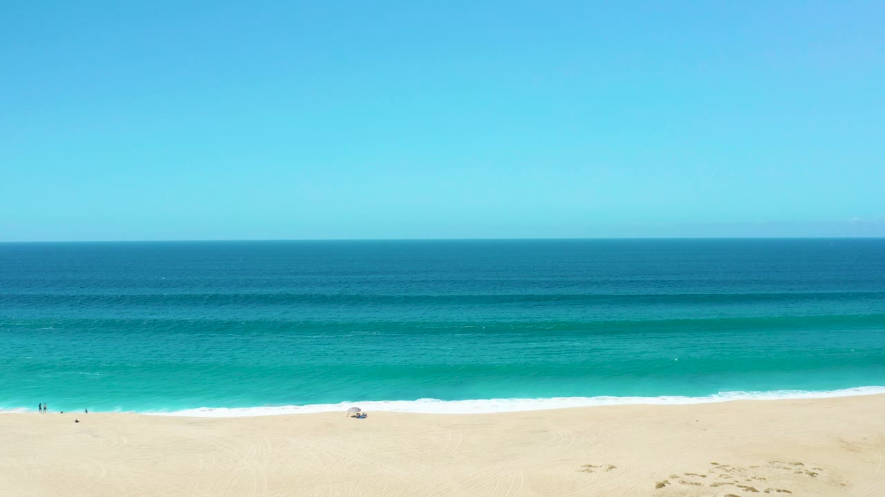 A wide La Laguna Beach Los Cabos frame shows deep blue water beyond turquoise shallows, white foam on the shore, and tiny people spaced along the sand.