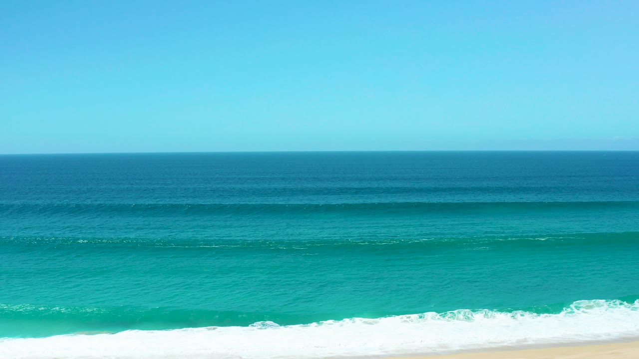 Turquoise ocean wave approaching a sandy beach under clear blue sky.