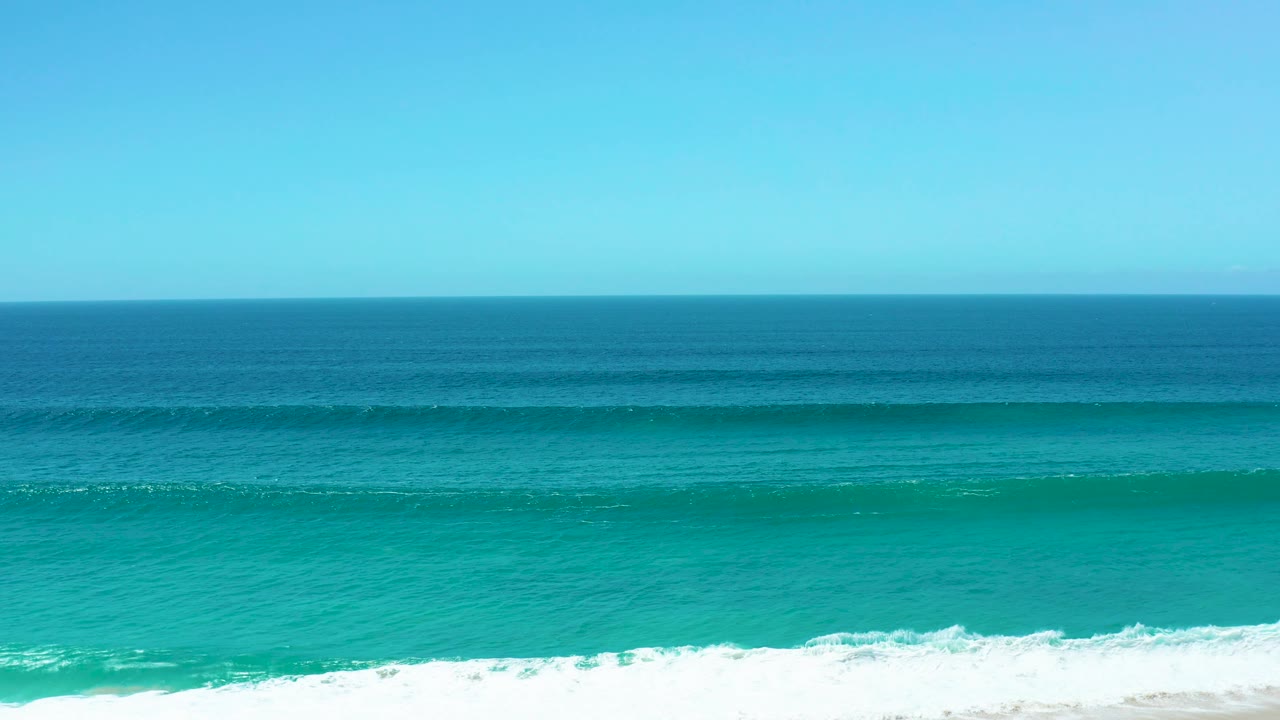 At La Laguna Beach Los Cabos, a blue-green wave band crosses the ocean surface while white foam collects along the sandy shoreline, with no visible people.