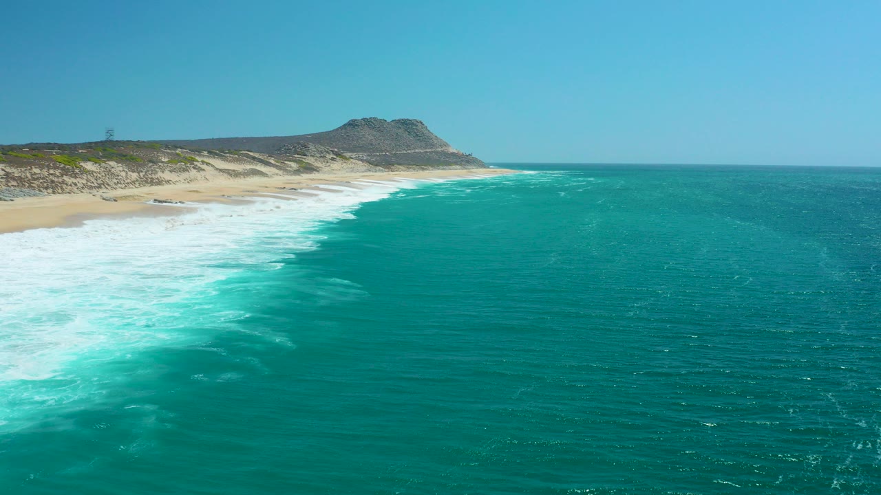 Aerial view at La Laguna Beach Los Cabos with turquoise ocean, white surf curving onto sand, low dunes, a distant hill, clear blue sky, and no visible people.