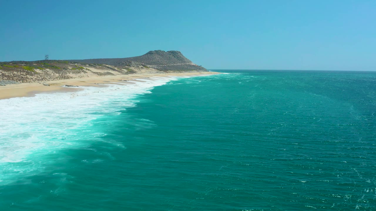 Wide aerial frame over La Laguna Beach Los Cabos with teal water across the foreground, white foam reaching the beach, dunes on the left, a hill beyond the shore, and no visible people.