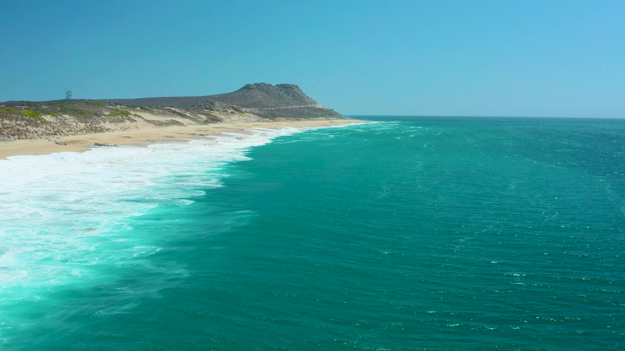 Foamy surf spreads across turquoise water beside a sandy shoreline.