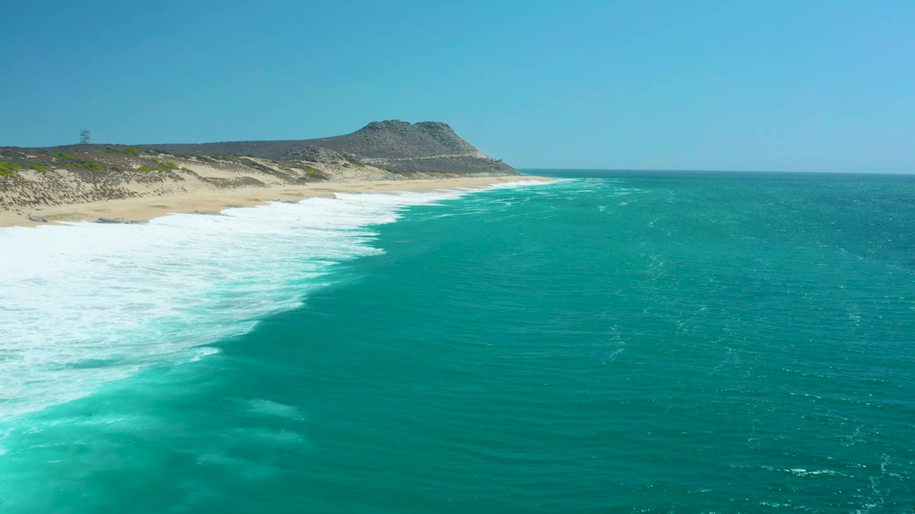 At La Laguna Beach Los Cabos, a broad band of white foam advances along the sandy shore while turquoise ocean fills the foreground, with no visible people.