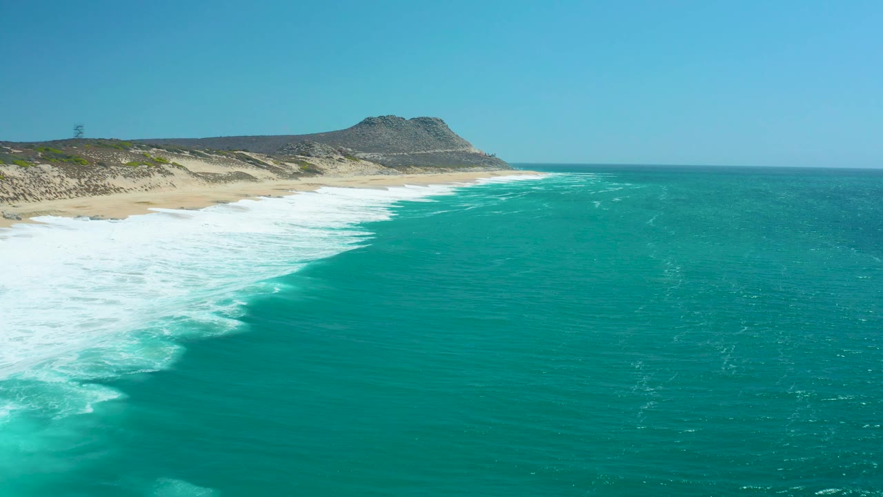 At La Laguna Beach Los Cabos, foamy surf spreads across the lower left shoreline while open turquoise water stretches to the right, with no visible people.