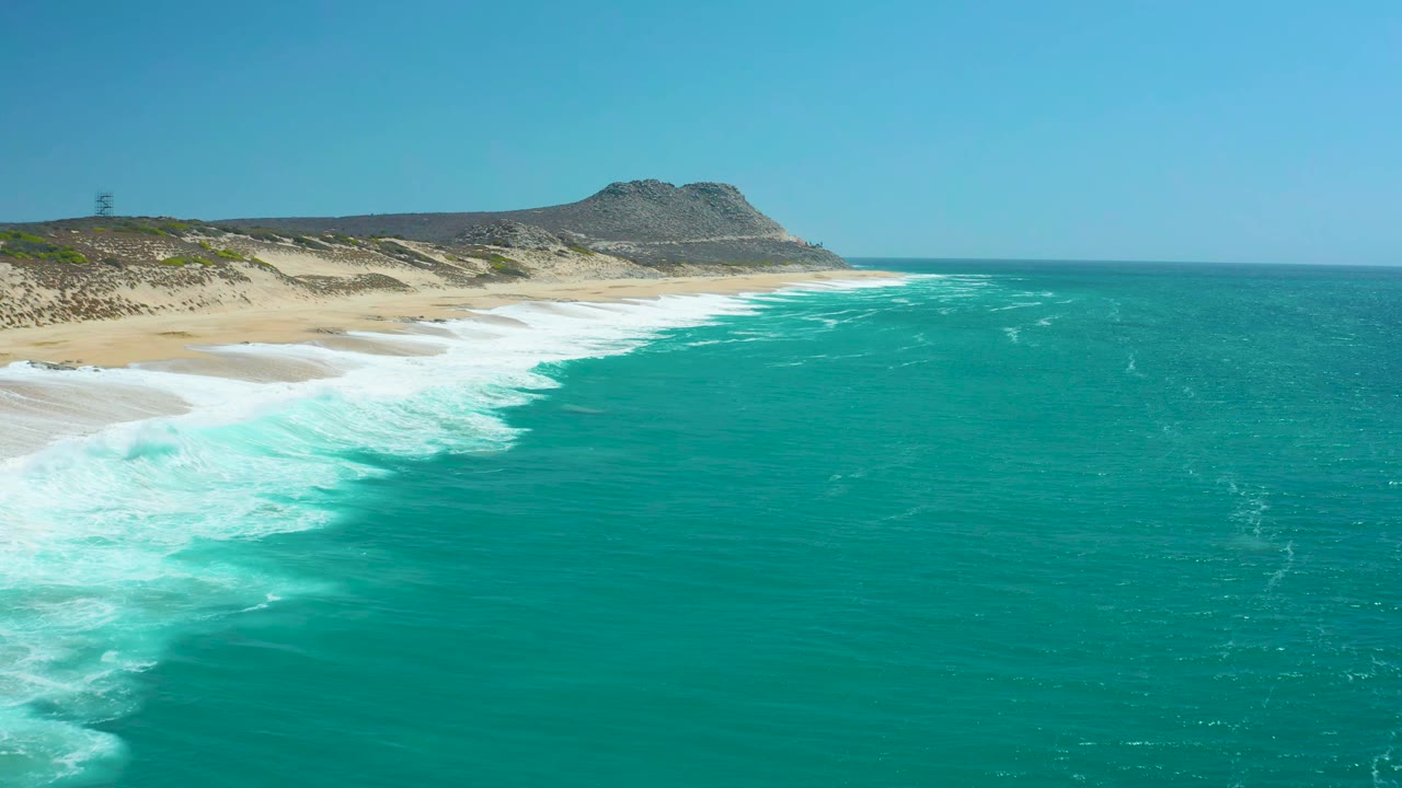 La Laguna Beach Los Cabos shoreline with the surf line pushed farther across the sand, turquoise ocean filling the right side, dunes and hill in back, and no visible people.
