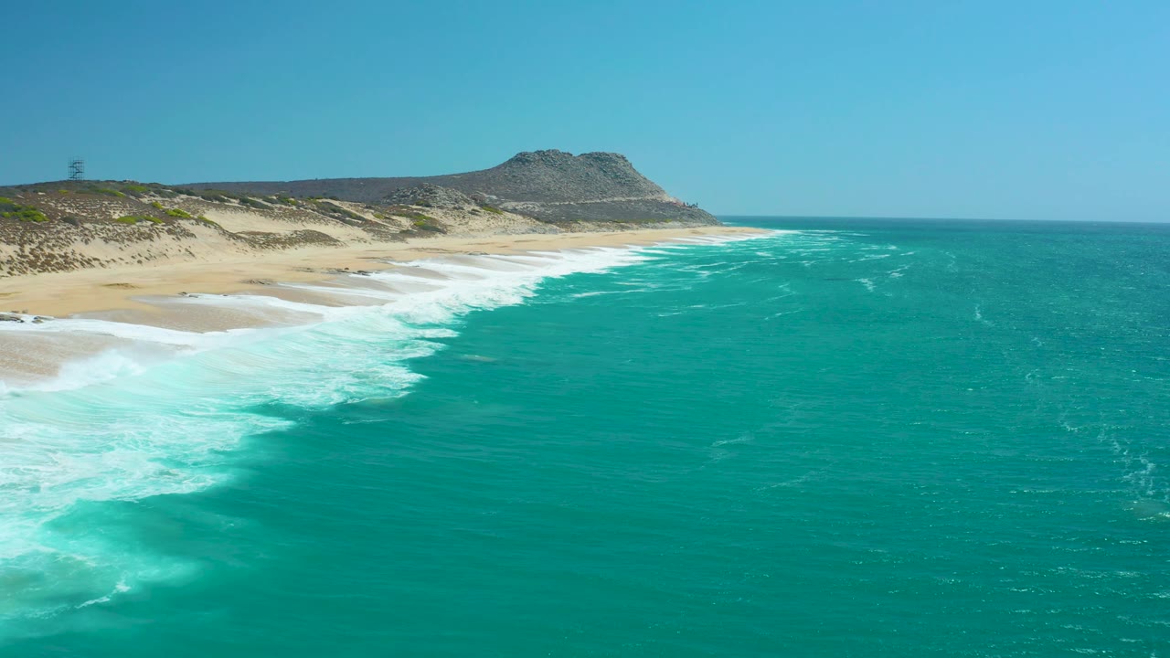 La Laguna Beach Los Cabos seen from above with more open water in the foreground, white waves tracking along the sand, dunes and a rocky hill beyond, and no visible people.