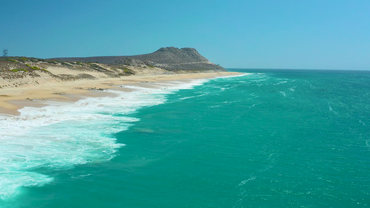 At La Laguna Beach Los Cabos, turquoise water pushes white surf onto a pale sandy shoreline with dunes and a low rocky hill under clear sky, no visible people.